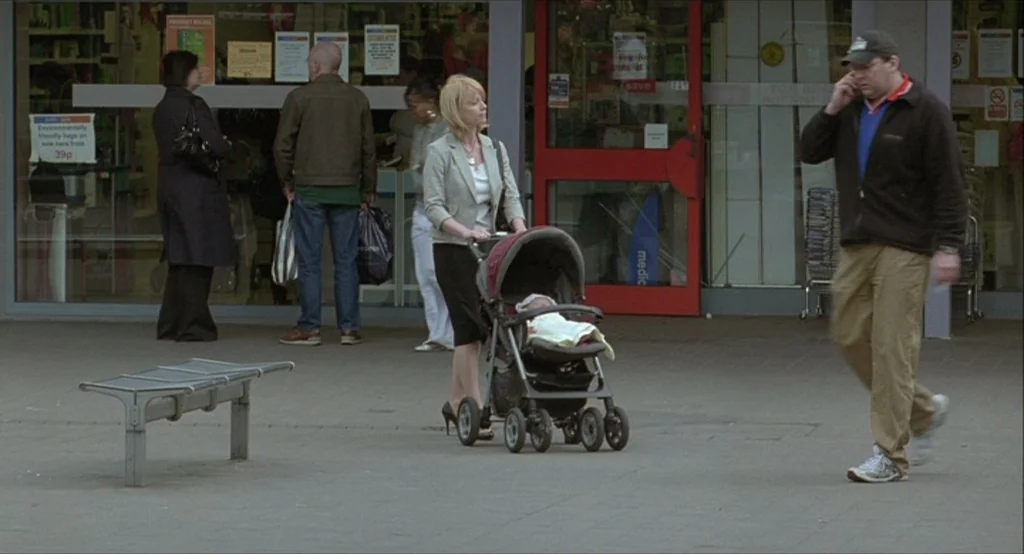 Looking for Eric (2009) movie still: fullbody — A commercial storefront with glass doors and advertisements sits behind a grey paved area. A woman…