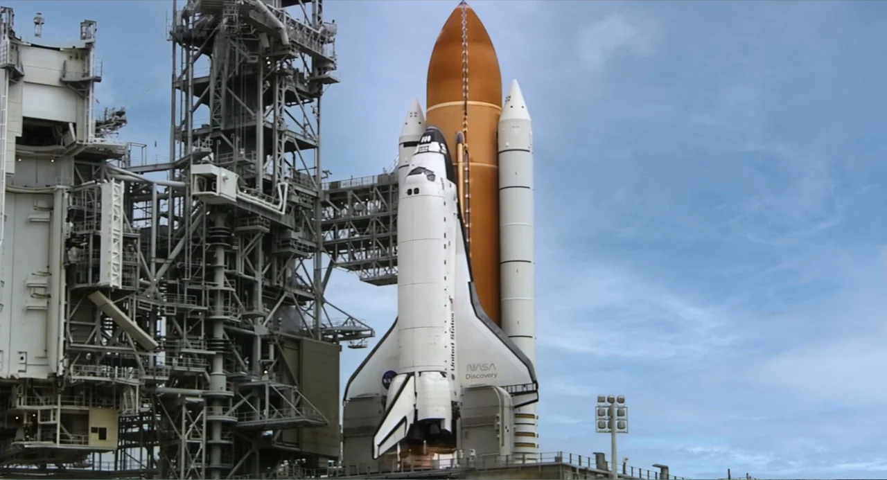 A Million Miles Away (2023) movie still: wide — A NASA space shuttle sits vertically on a massive metal launchpad structure under daylight. The…