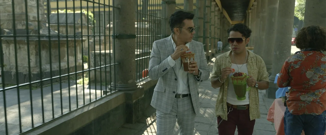 Divine Intervention (2026) movie still: medium — A stone colonnade lined with metal fencing overlooks an older architectural site with statues. Two…