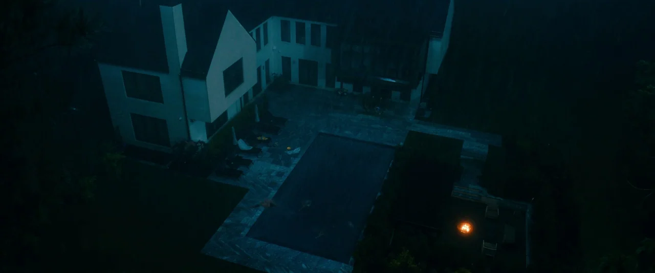 Leave The World Behind (2023) movie still: establishing — A modern two-story house sits by a rectangular pool during a heavy rainstorm. A small fire…