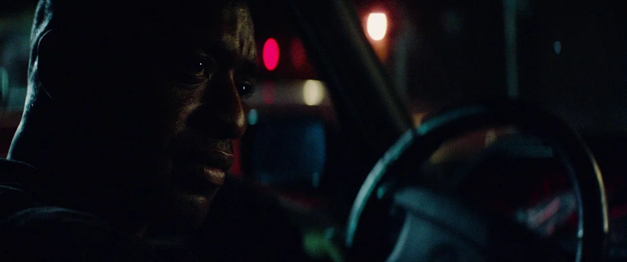 Triple 9 (2016) movie still: closeup — The interior of a vehicle at night with blurred city lights visible outside. A man sits in the driver seat…