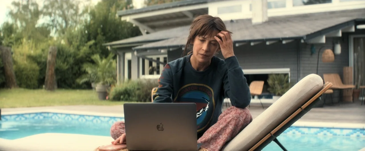 I Love America (2022) movie still: medium — A woman sits on a patio lounge chair beside a swimming pool outside a house. She uses a laptop computer…