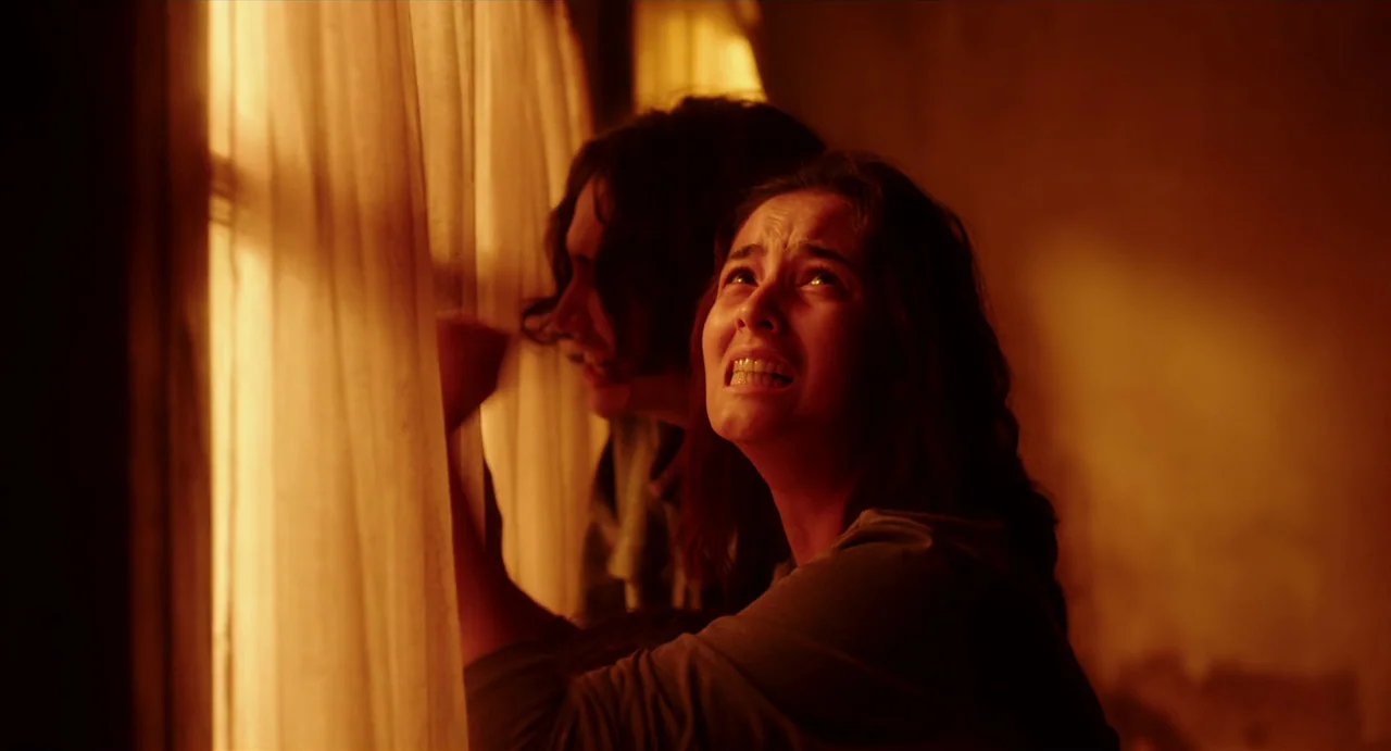 May The Devil Take You (2018) movie still: medium — A darkened interior space with a single window covered by light fabric curtains. Two people…