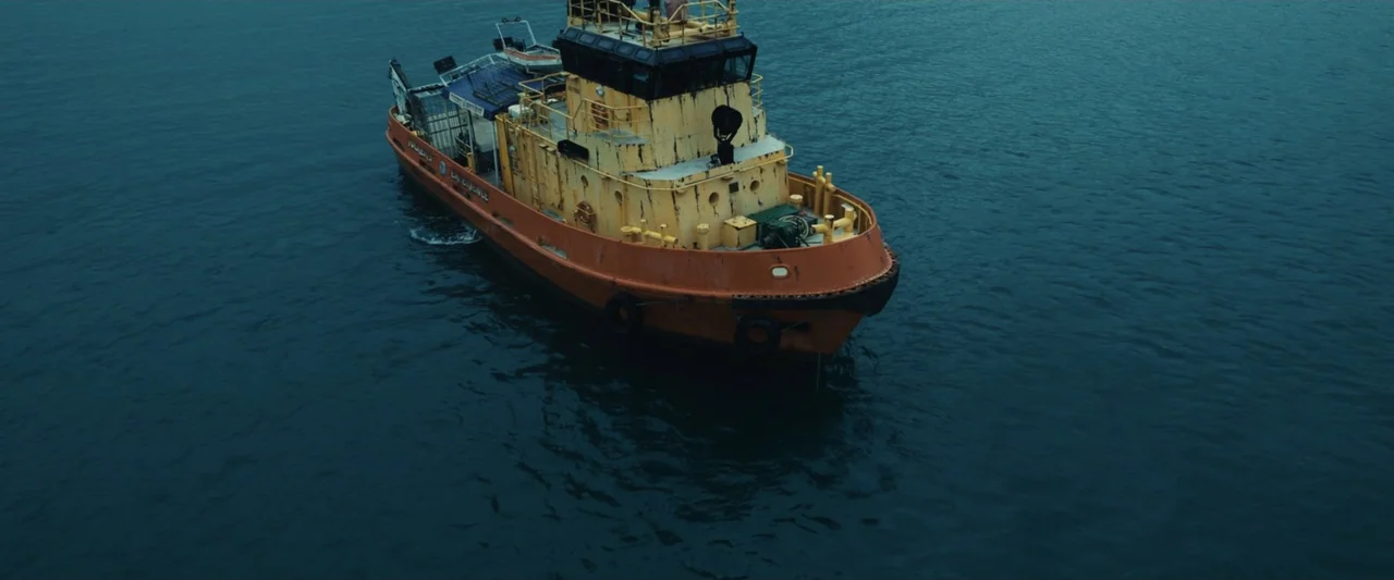 Dangerous Animals (2025) movie still: wide — A weather-worn orange and yellow tugboat sits on calm, deep blue open water. The vessel shows signs of…