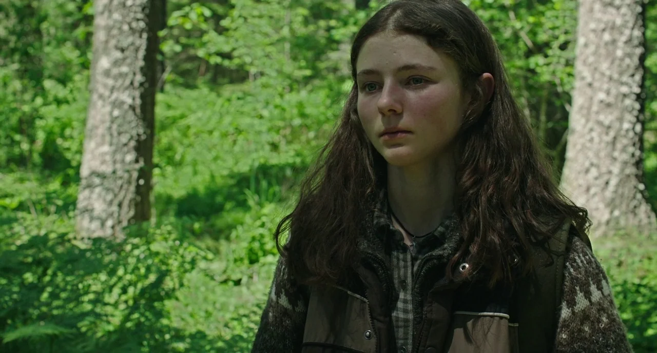 Leave No Trace (2018) movie still: medium — A teenage girl stands in the middle of a dense, lush green woodland area. She looks forward with a…