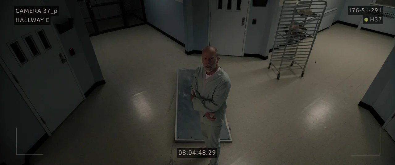 Glass (2019) movie still: wide — A sterile, brightly lit facility room features tile flooring and white security doors. An elderly man in medical…