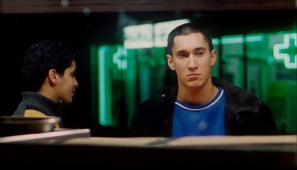 Nenette et Boni (1996) movie still: medium — A street corner at night illuminated by the green glow of a pharmacy cross sign. Two men face each…