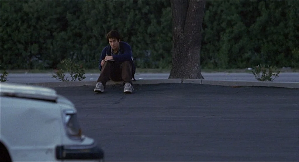 The Good Girl (2002) movie still: wide — An asphalt parking lot borders a manicured green hedge and a single mature tree. A man sits on the concrete…
