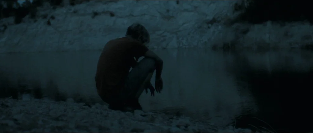 Lasting (Nieulotne) (2013) movie still: medium — A dark riverbank at night with a rocky edge meets a calm, murky stream. A person is crouched near…