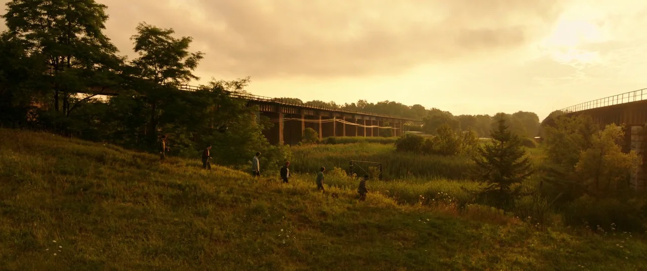It Chapter Two (2019) movie still: wide — A group of six teenagers walks across an overgrown field during golden hour. A long wooden railroad…