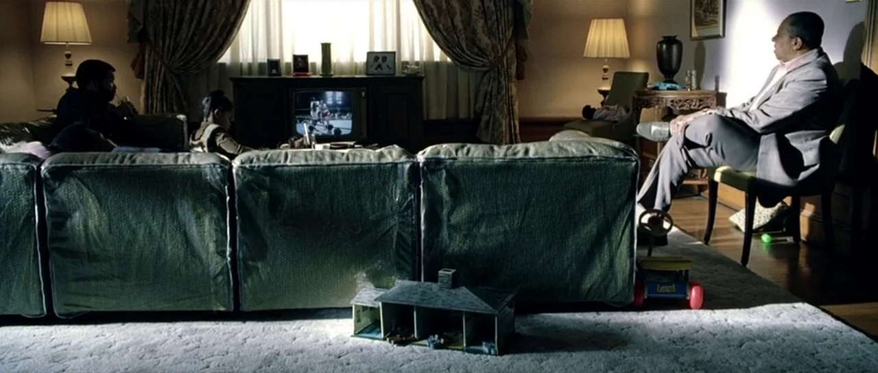 Ali (2001) movie still: wide — A dark living room contains a large couch covered in transparent plastic protective sheets. Several individuals watch…