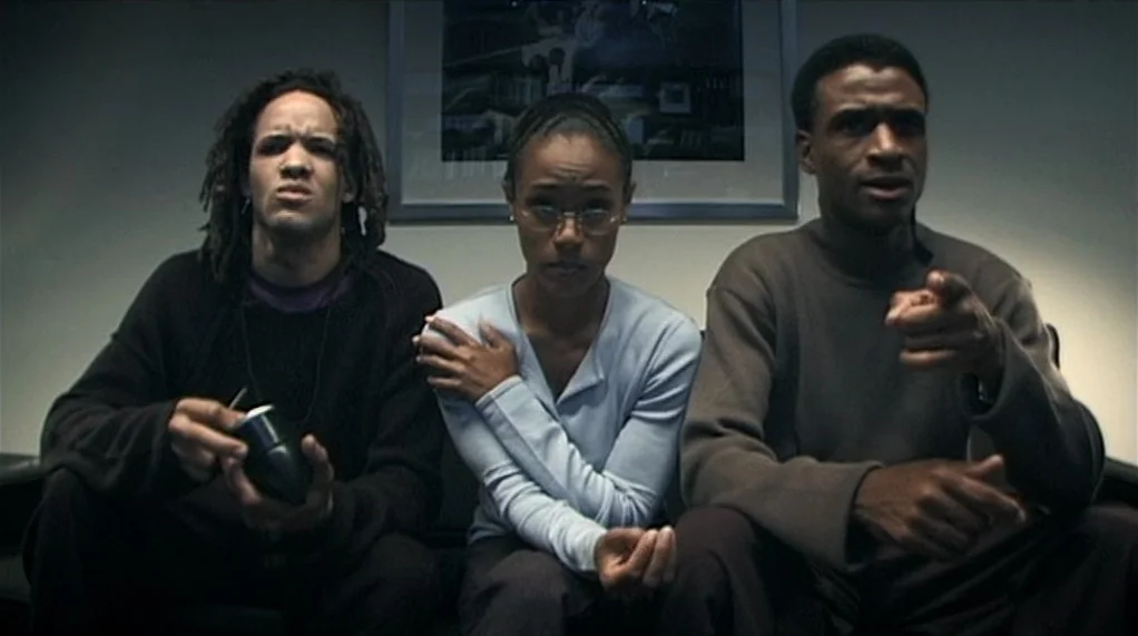 Bamboozled (2000) movie still: medium — Three young adults sit on a couch against a plain grey wall with framed art. The man on the right points his…