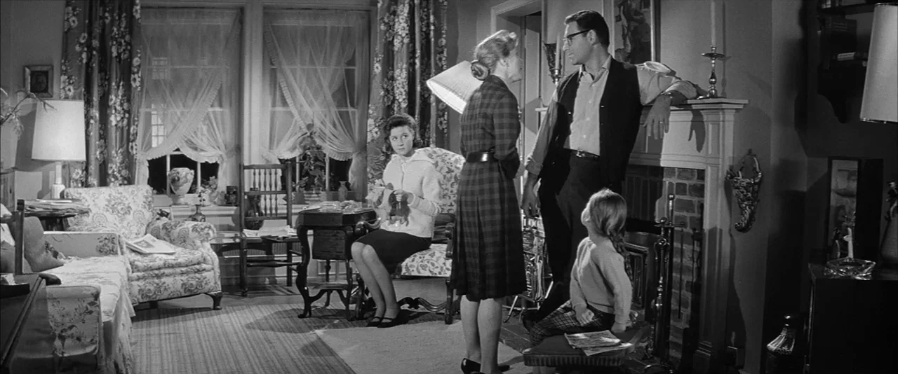 The Haunting (1963) movie still: wide — A 1950s suburban home interior features floral curtains, patterned armchairs, and a brick fireplace. A man…