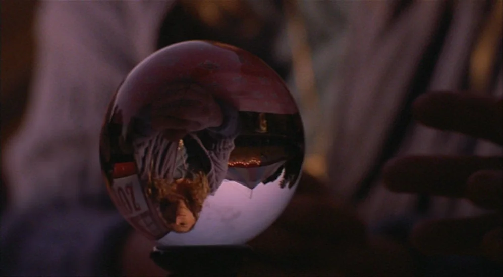 Roselyne and the Lions (1989) movie still: closeup — A dark outdoor space features a glass crystal ball resting on a stand. The ball reflects an…