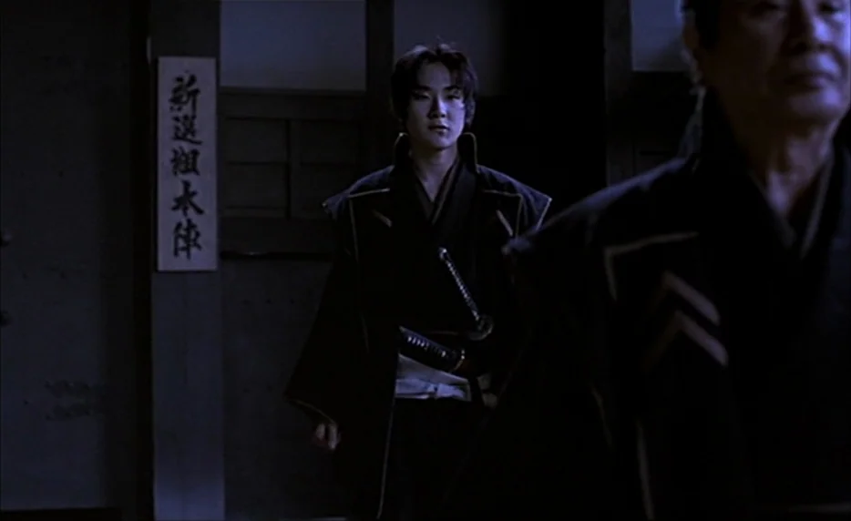 Gohatto (1999) movie still: medium — A dimly lit indoor hallway with a wooden wall featuring a vertical white sign. A young man wearing a katana…