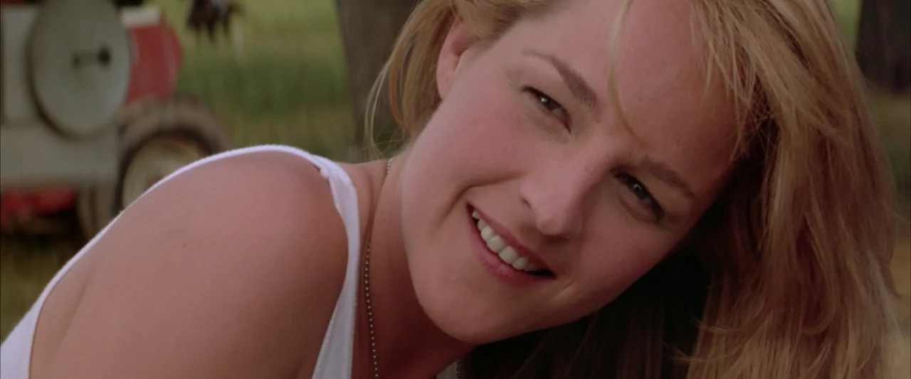 Twister (1996) movie still: closeup — An outdoor field setting with a blurred agricultural vehicle in the distance. A woman looks at the camera…