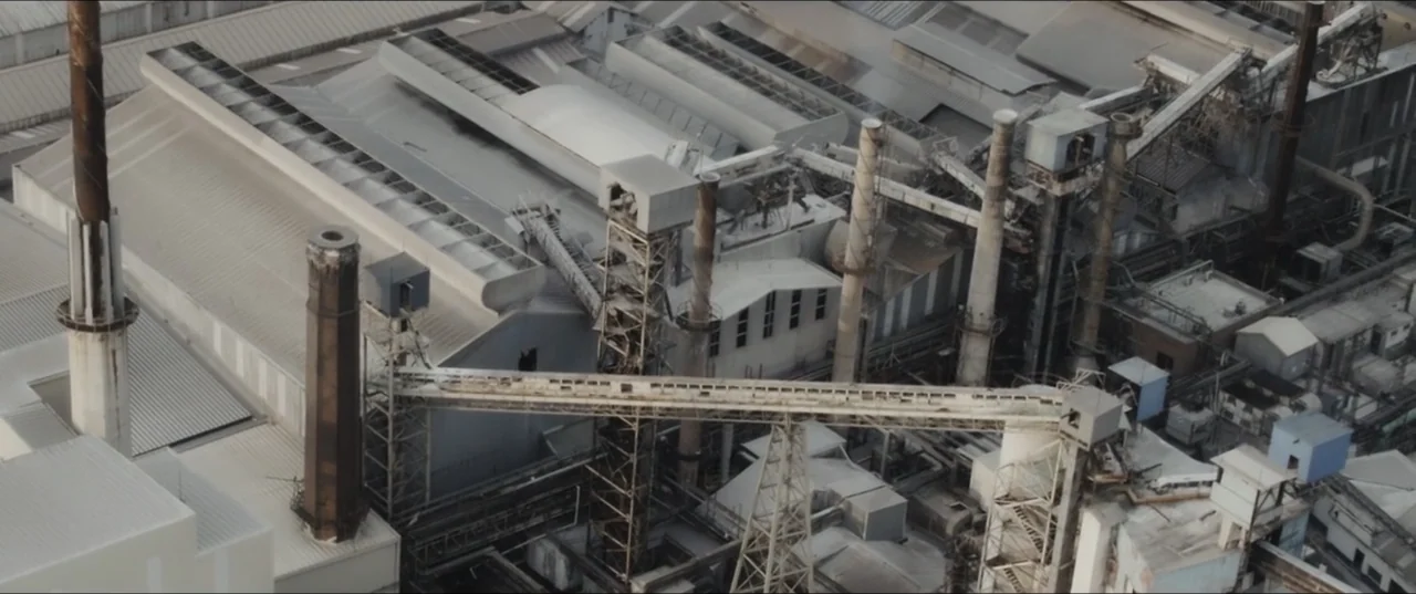 Death to Summer (2021) movie still: establishing — An overhead view of a large industrial complex featuring multiple metal chimneys and piping. The…