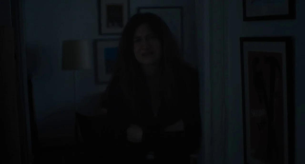 Private Life (2018) movie still: medium — A dimly lit hallway features framed prints on the walls in low light. A woman with long hair stands near a…