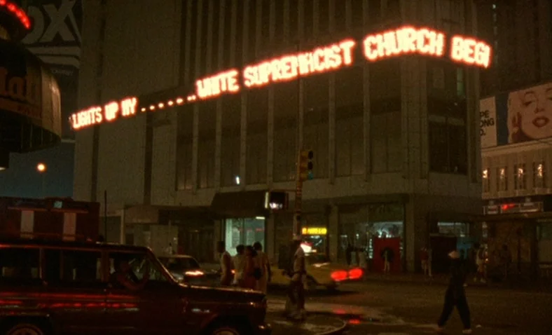 Paris Is Burning (1990) movie still: wide — A dark city intersection features a large, glowing red LED sign suspended across the street. The sign…