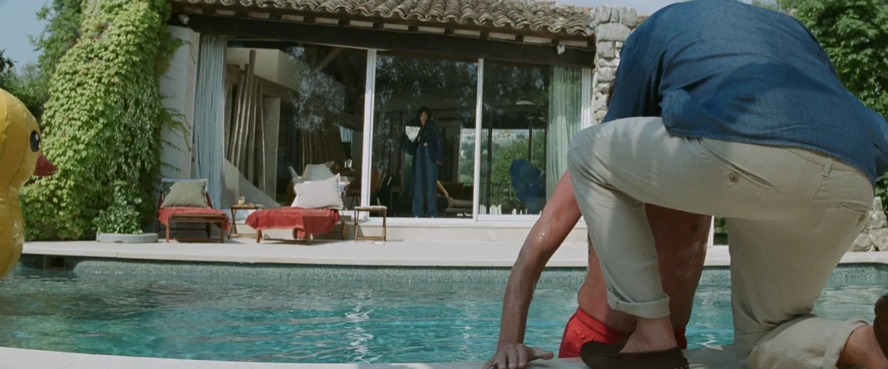 Masquerade (2022) movie still: medium — A modern Mediterranean villa with a glass-walled living room overlooks a tiled swimming pool. A man in a…