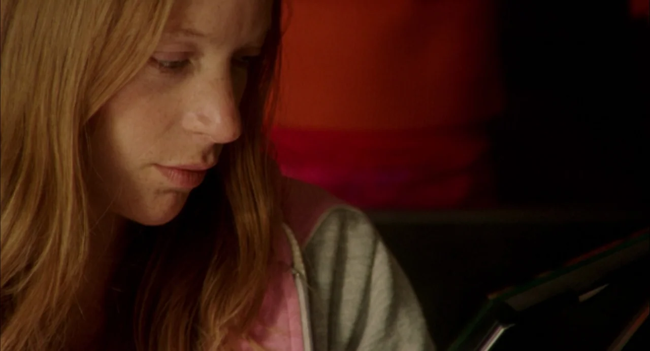 My Summer of Love movie still: closeup — A woman looks down at an item in a dimly lit indoor space. She wears a pink hoodie while focused on her task.