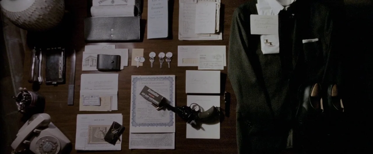 A Single Man (2009) movie still: establishing — A dark wooden desk surface is organized with various legal papers, personal items, and clothing. A…