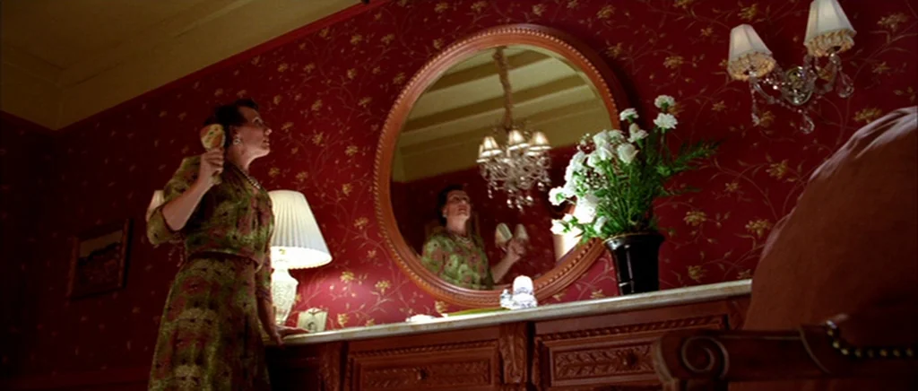 The Aviator (2004) movie still: medium — A woman stands before a large round mirror in a room with deep red wallpaper. She holds a hairbrush and…