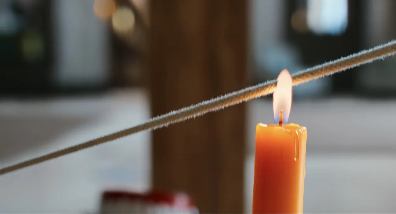 Tom & Jerry (2021) movie still: closeup — A burning orange candle is positioned in the foreground of a blurred interior room. The flame touches a…