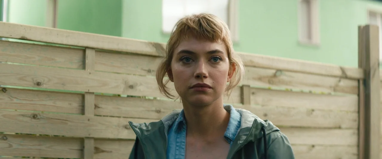 Vivarium (2019) movie still: closeup — A woman stands in front of a wooden horizontal-slat fence outdoors. A green building is visible in the…