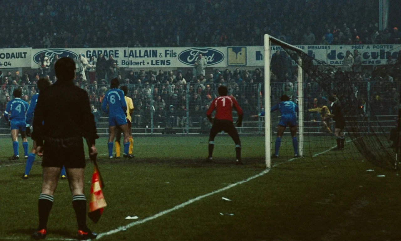 Graduate First (1978) movie still: wide — A football match takes place at night in a crowded, fenced stadium. Players in blue and red jerseys line…