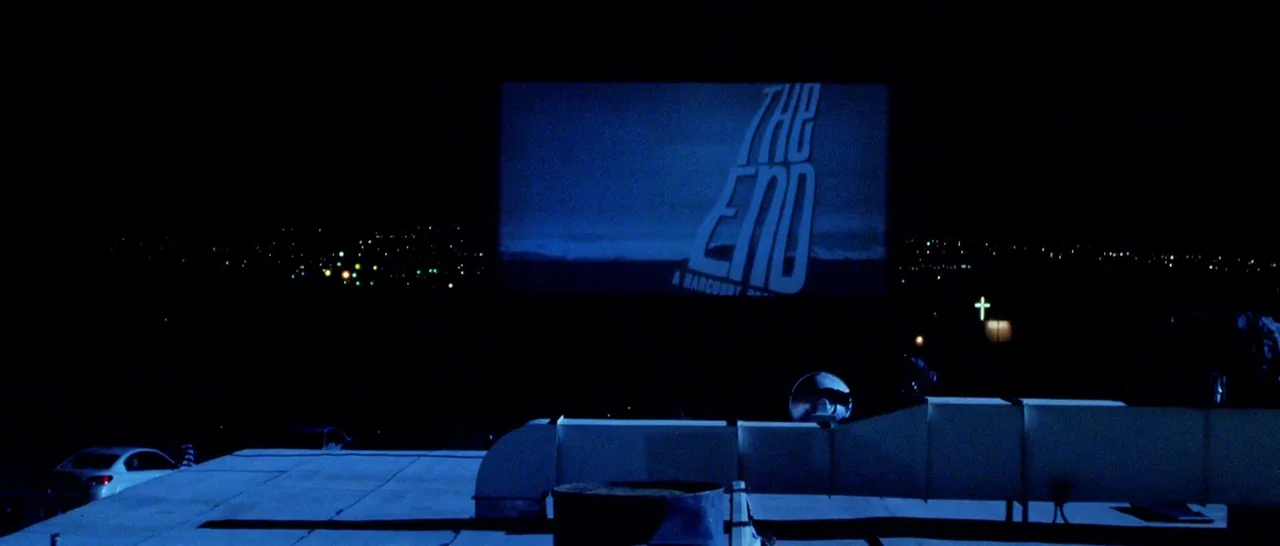 Too Late (2015) movie still: wide — A rooftop view overlooks a drive-in theater screen at night. The screen features a film title card displaying…