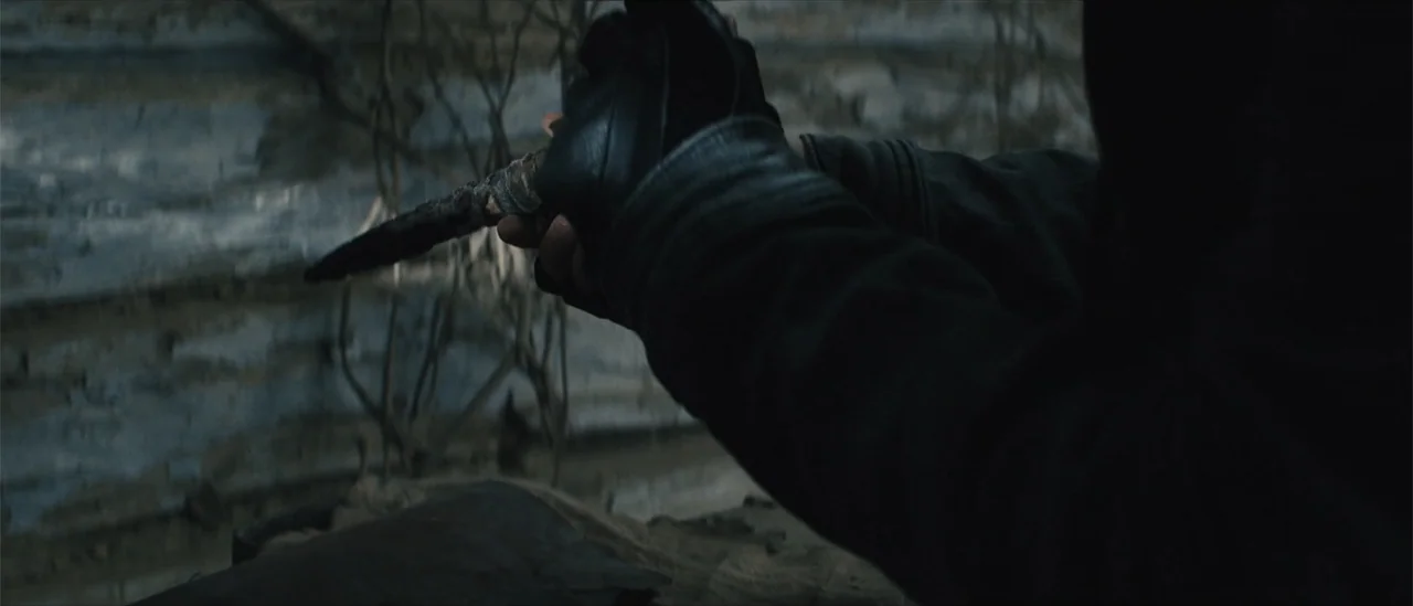 All Fun and Games (2023) movie still: closeup — A dark, weathered environment with textured debris and dead branches in the background. A person's…