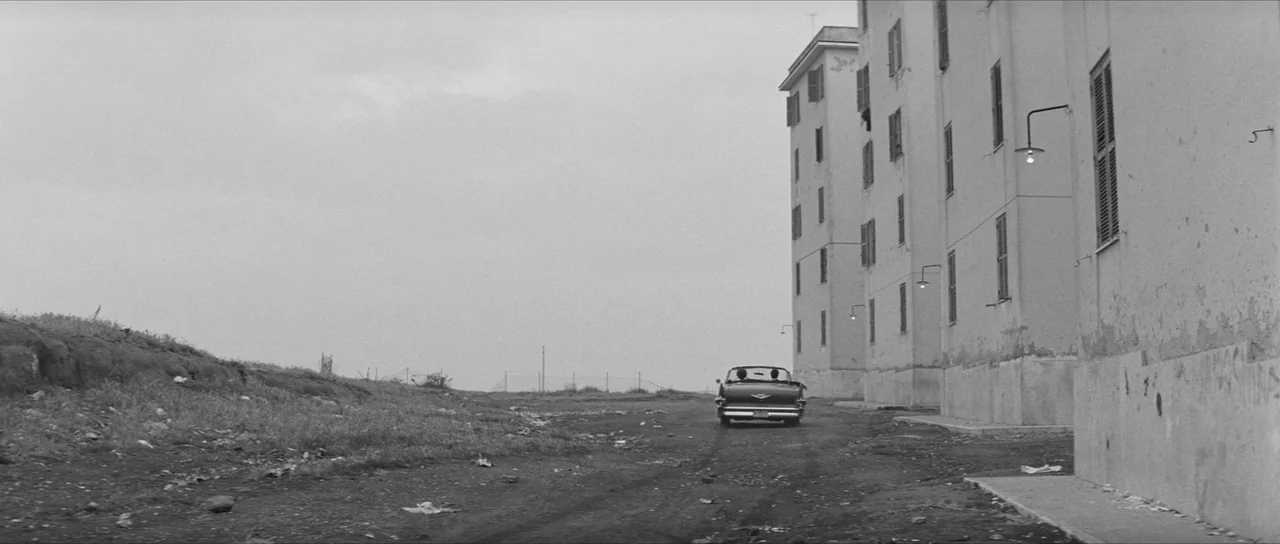 La Dolce Vita (1960) movie still: wide — A dark convertible drives along an unpaved road adjacent to a tall, plain apartment building. The area is…