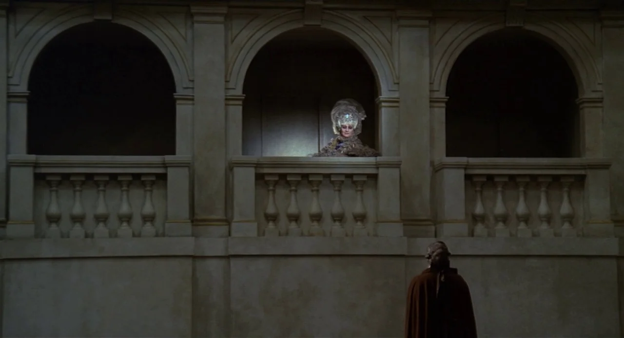 Fellini’s Casanova (1976) movie still: wide — A stone balcony with three arched openings overlooks a dim theater space. A woman in an elaborate…
