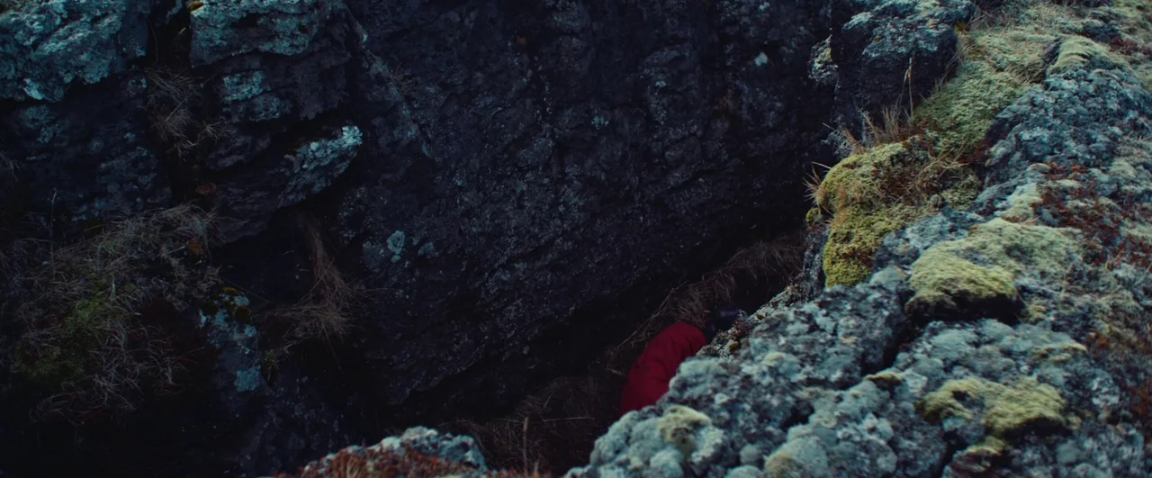 Rift (2017) movie still: wide — A deep, narrow fissure in volcanic rock is lined with patches of green moss. A figure wearing a red jacket rests on…