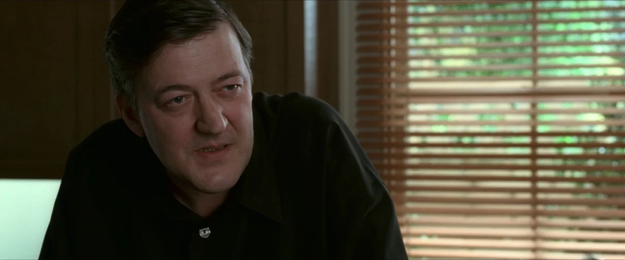 V for Vendetta (2006) movie still: medium — A man sits in an interior space near a window with wooden blinds. The man wears a black collared shirt…