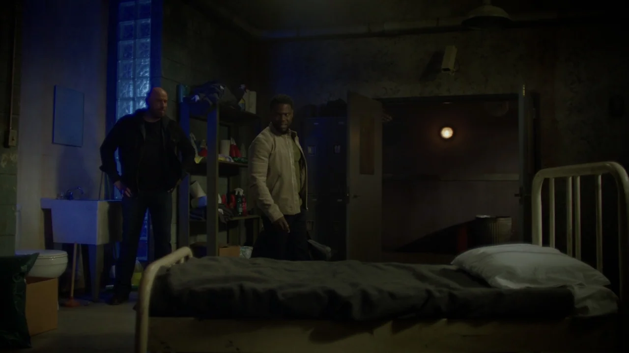 Die Hart (2023) movie still: fullbody — A dimly lit room with grey concrete walls features a metal bed frame and shelving. Two men stand in the…