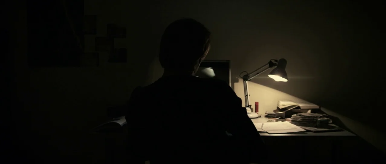 Vanishing Waves (2012) movie still: medium — A dark space features a person sitting at a small table at night. The table holds an open lamp, papers,…