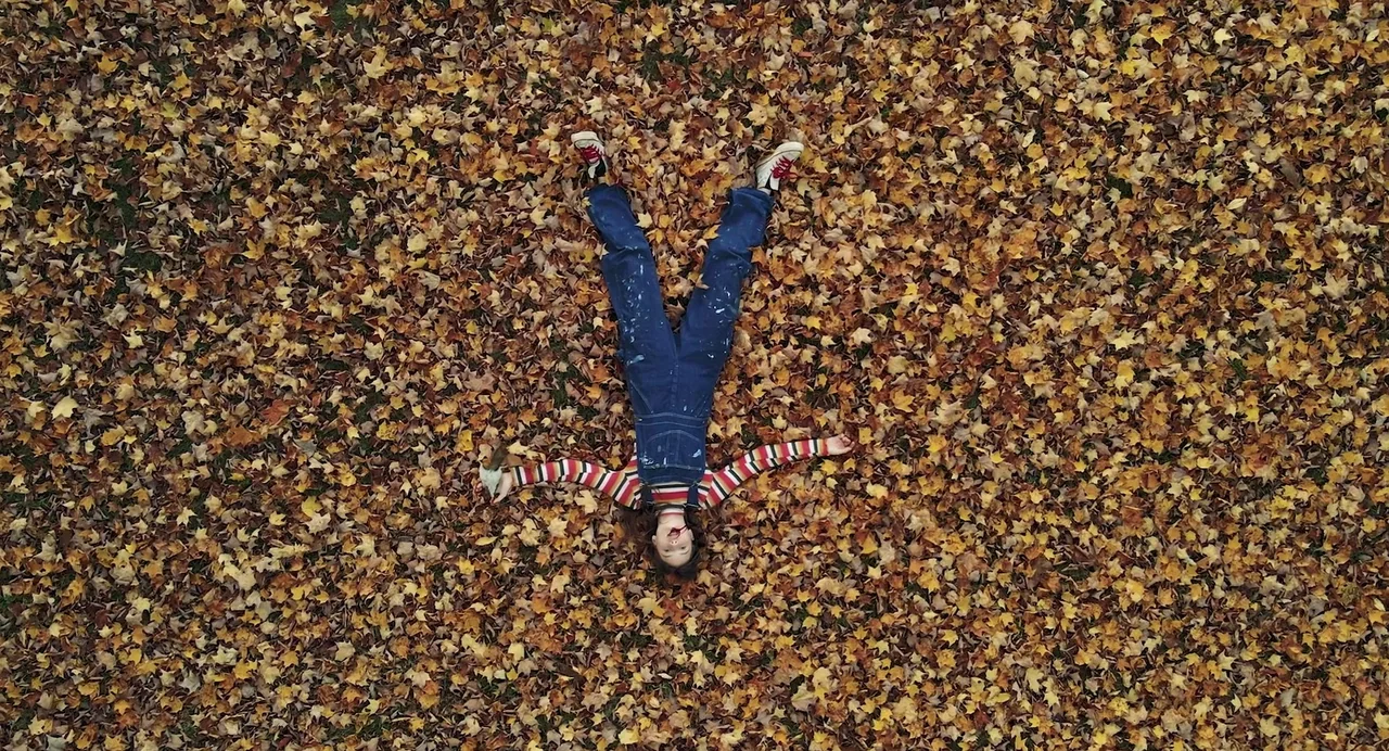 Hellbender (2021) movie still: fullbody — A person lies amidst a thick blanket of fallen autumn leaves on the ground. They are positioned in the…