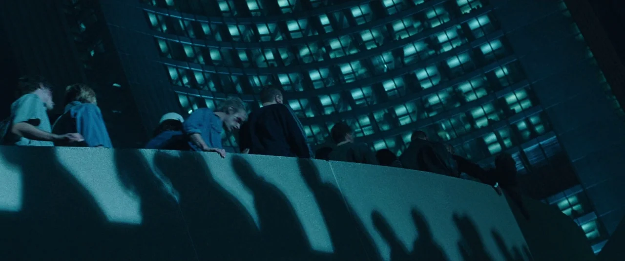 Resident Evil: Apocalypse (2004) movie still: wide — A modern curved building facade glows with grid-like teal windows at night. A group of people…