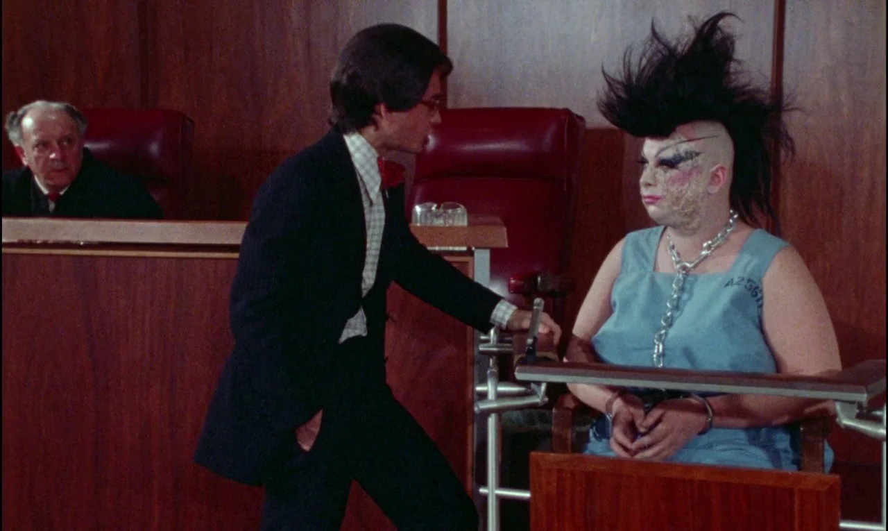 Female Trouble (1974) movie still: medium — A wood-paneled room features a judge's bench and a prisoner seated at a table. A man in a suit speaks to…