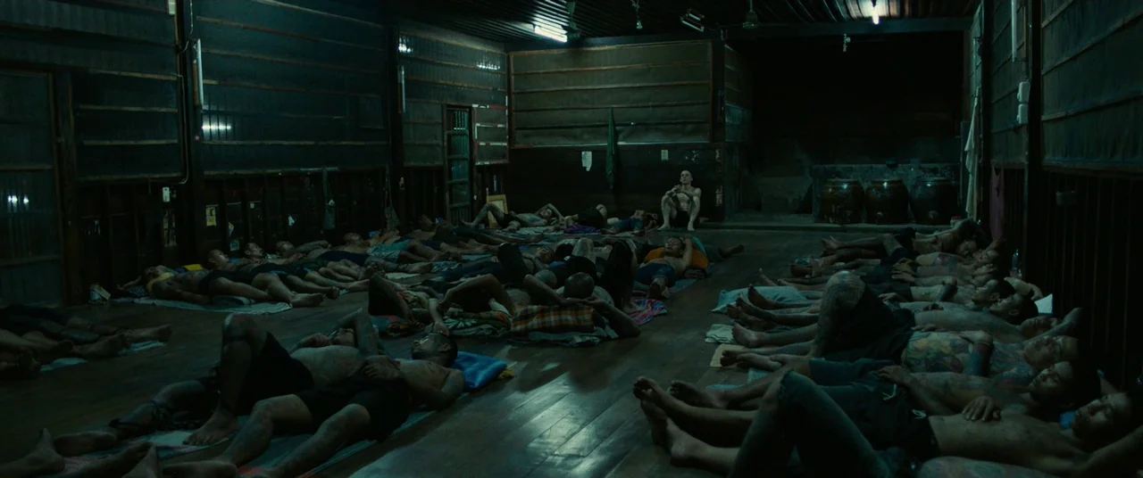 A Prayer Before Dawn (2017) movie still: wide — A dark wooden dormitory houses many men sleeping closely together on thin floor mats. One lone man…