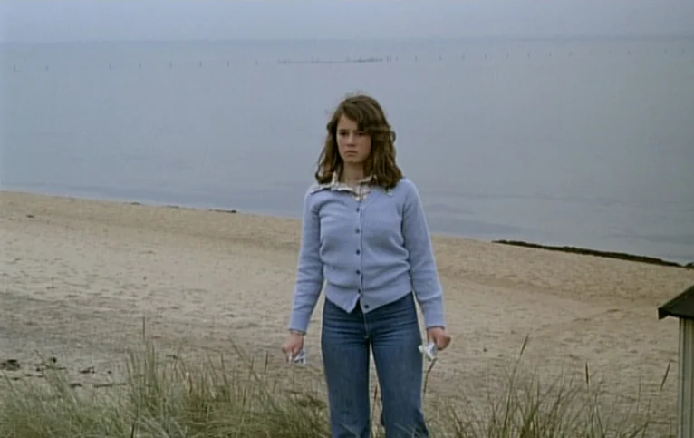 Giliap (1975) movie still: fullbody — An overcast beach landscape features tan sand and a calm body of water. A teenage girl stands in the grass…