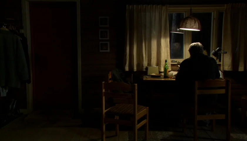 Saraband (2003) movie still: wide — A dimly lit room with wood-paneled walls features a dining area by a window. A man sits alone at the table under…