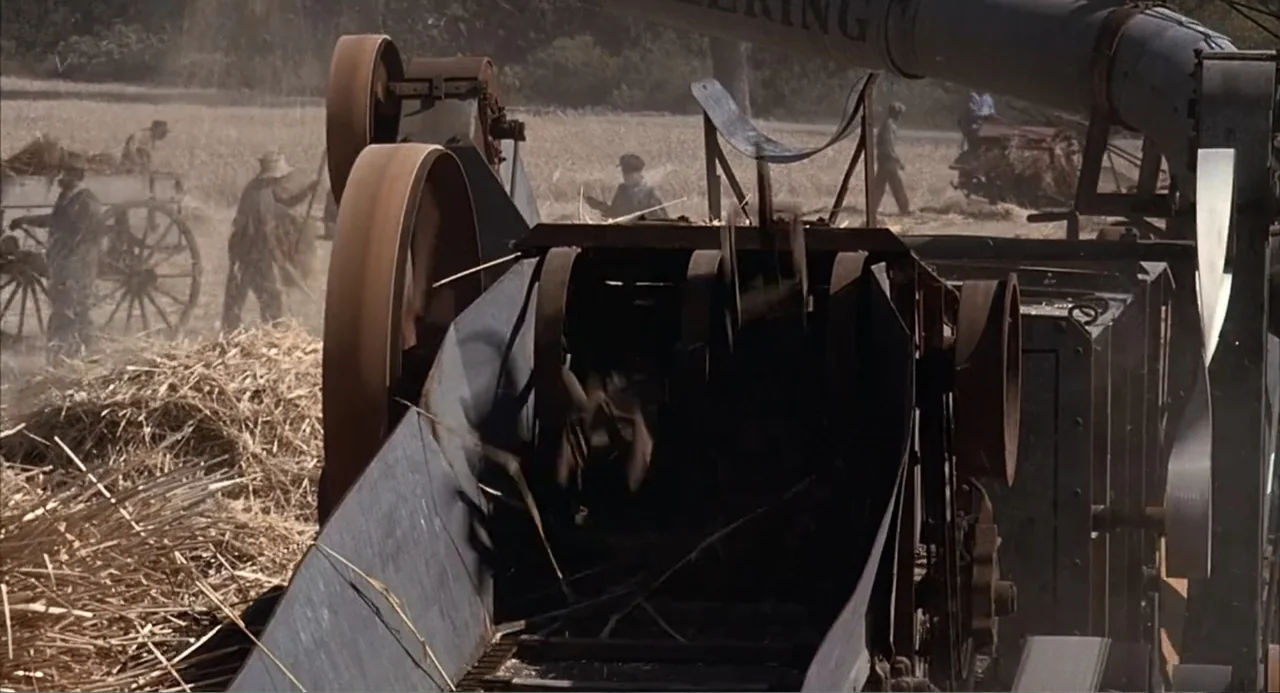 Of Mice and Men (1992) movie still: medium — An industrial threshing machine processes harvested grain in a dusty rural agricultural field. Several…