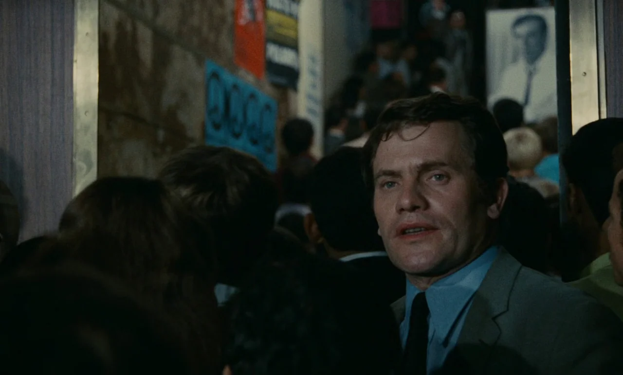 Z (1969) movie still: medium — A crowded subway carriage features posters on the wall and many standing commuters. A man in a grey suit and blue…
