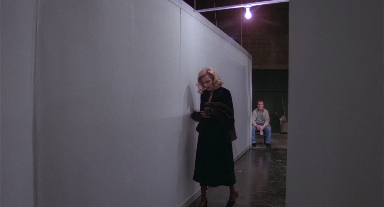 Opening Night (1977) movie still: fullbody — A stark, minimalist white partition wall stands in a dark, unfinished-looking interior space. A blonde…