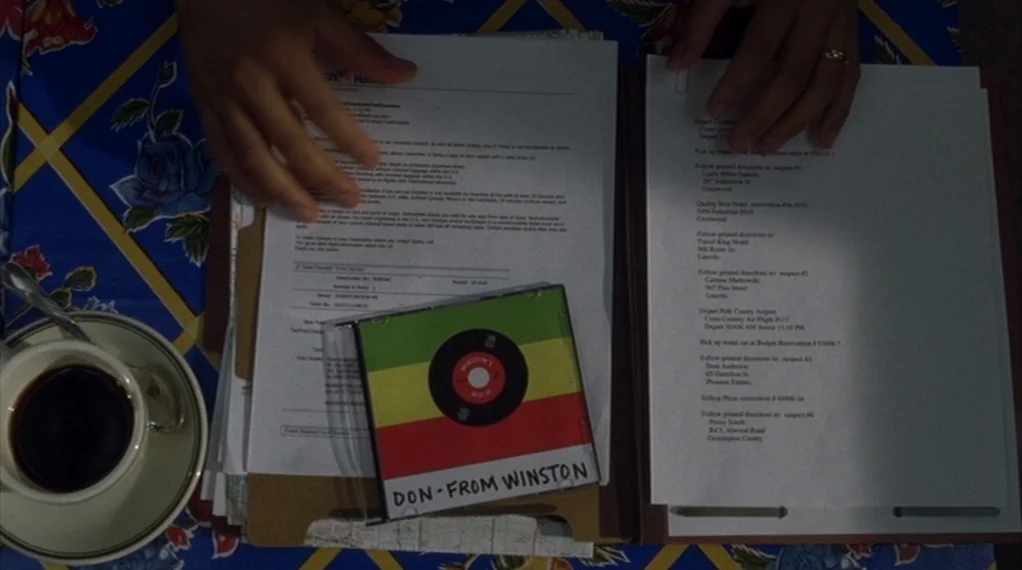 Broken Flowers movie still: closeup — A top-down view shows printed documents and a music disc on a patterned tablecloth. A white ceramic coffee cup…