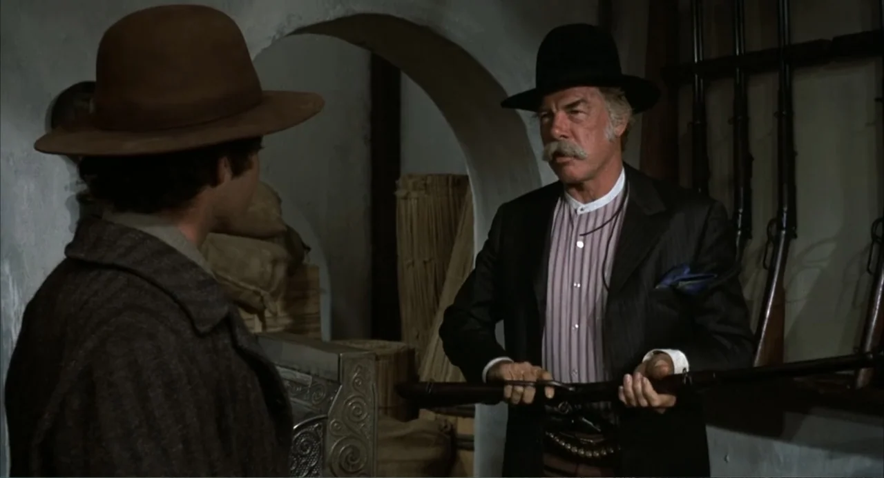 The Spikes Gang (1974) movie still: medium — A dimly lit room with an arched doorway and several rifles mounted on walls. An older man holds a rifle…