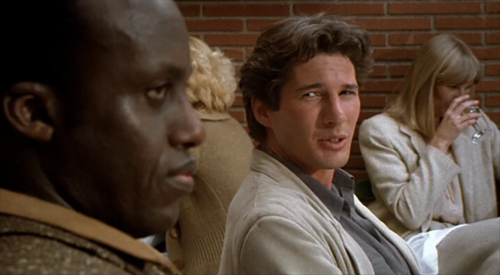 American Gigolo (1980) movie still: medium — A group of people sit in a space with a reddish-brown brick wall. The man in the center speaks while a…